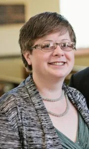 A smiling person with short hair and glasses, perhaps one of the renowned world literature speakers, wears a patterned gray sweater over a teal top and a beaded necklace. They are indoors with a blurred background.