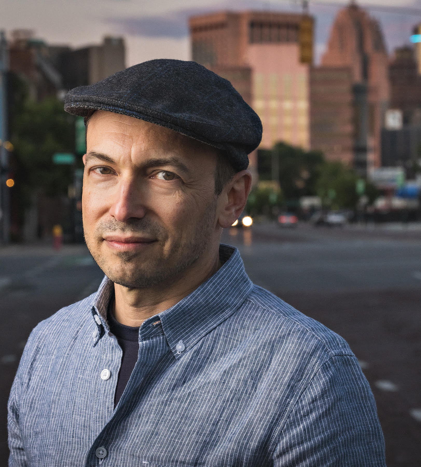A person wearing a flat cap and a striped button-up shirt stands on a city street with buildings in the background during twilight.
