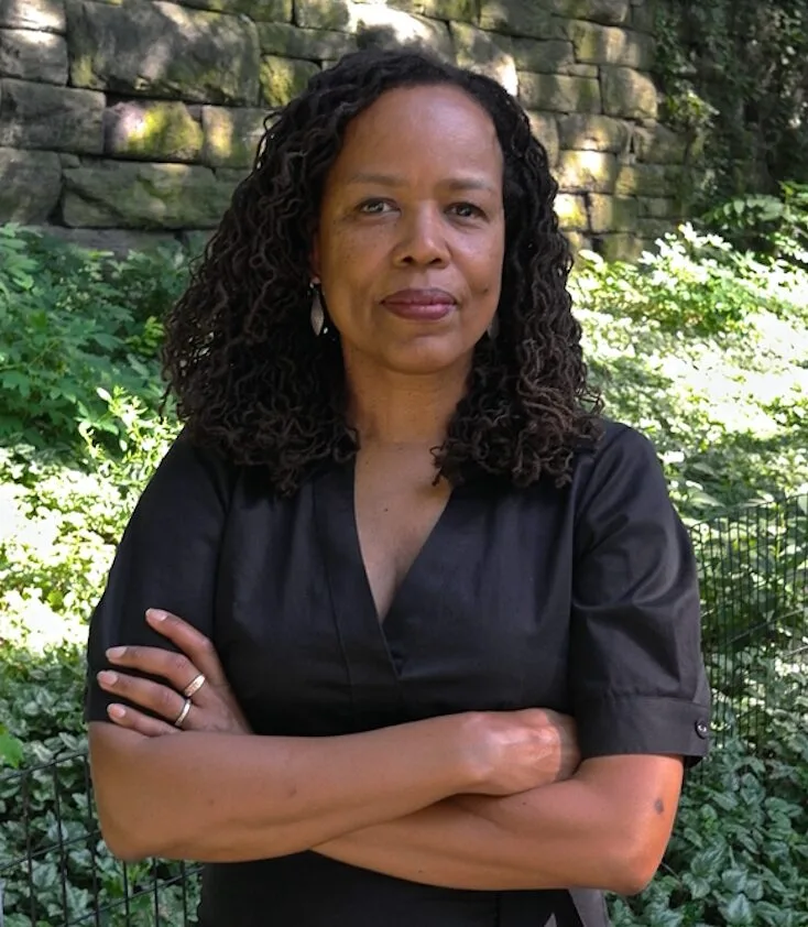 A woman with curly hair stands outdoors in front of a stone wall and greenery, wearing a black short-sleeve dress with her arms crossed, looking confidently at the camera.
