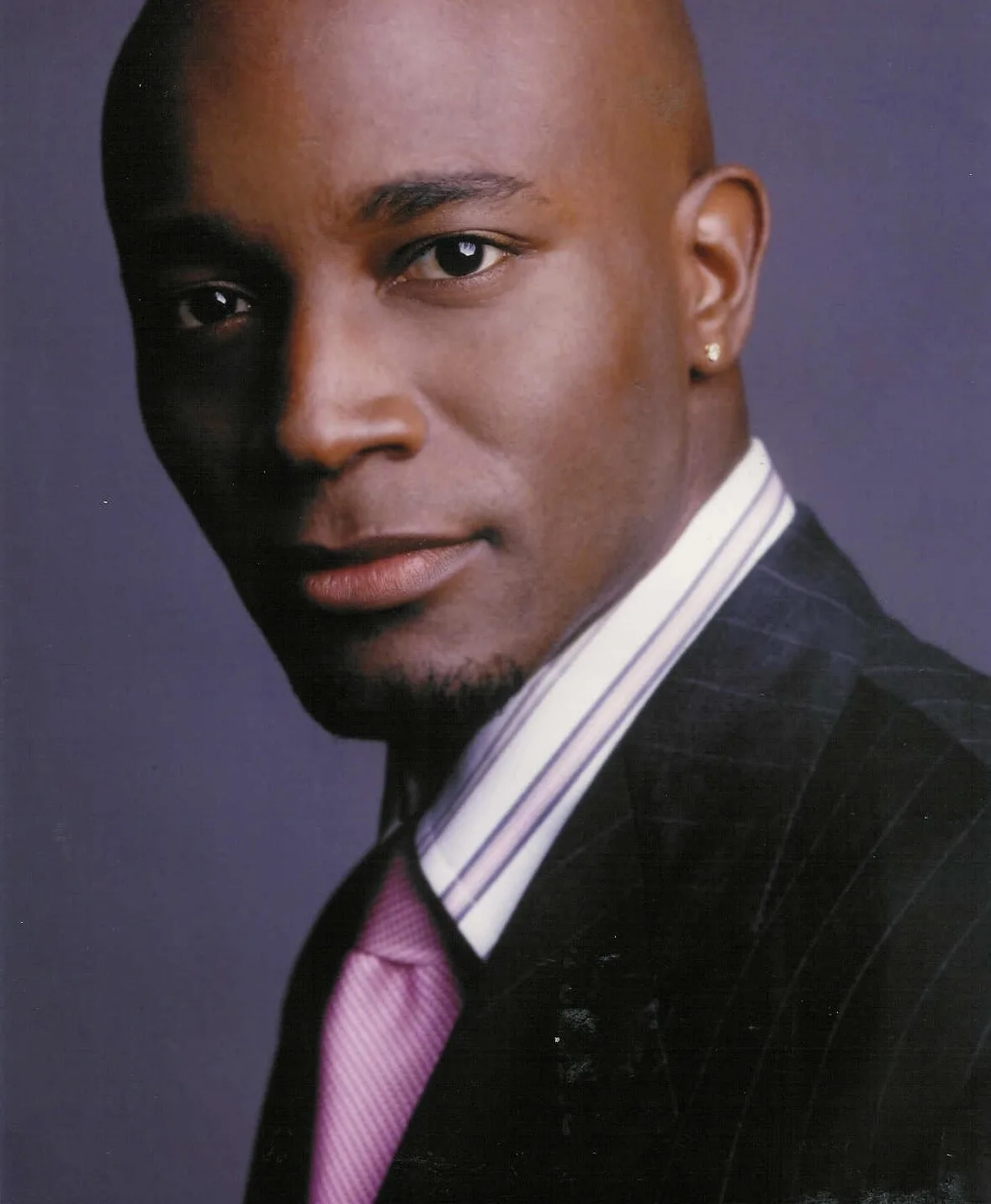 A person with a shaved head and an earring in their left ear is wearing a pinstripe suit, a white shirt, and a pink tie, posed against a smooth purple background.