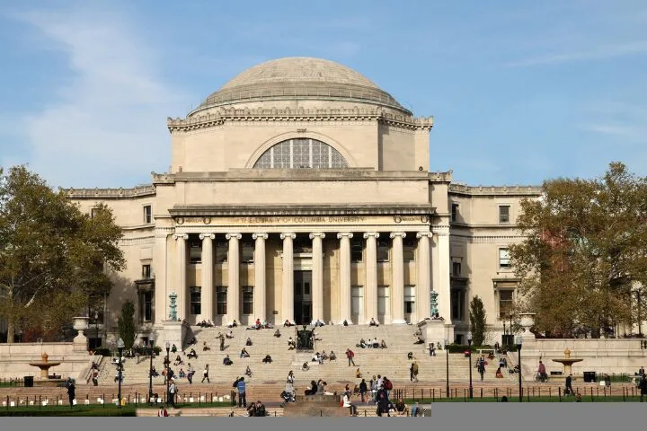 A large neoclassical building with a domed roof and tall columns, surrounded by trees. Many people are sitting or walking on the wide steps and plaza in front of the building under a clear sky.
