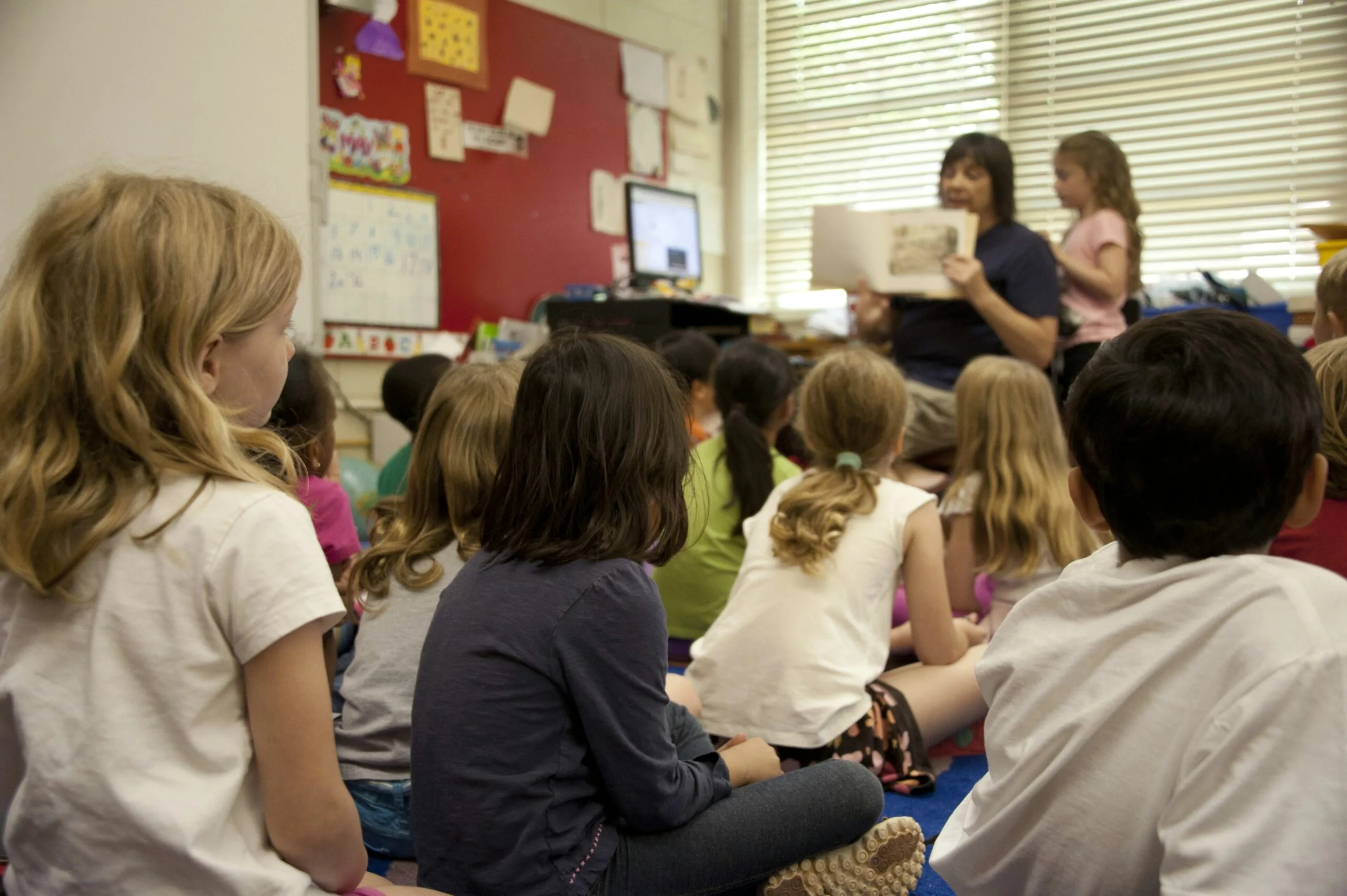 A group of young children sit on the floor, attentively listening to an adult reading a book at the front of a classroom. The room is colorful, with educational posters on the walls and sunlight filtering through blinds.