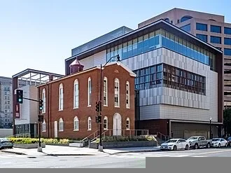 A historic brick church building stands in front of a large, modern glass and metal structure at a city street corner, with cars parked along the curb and a few traffic signals visible.