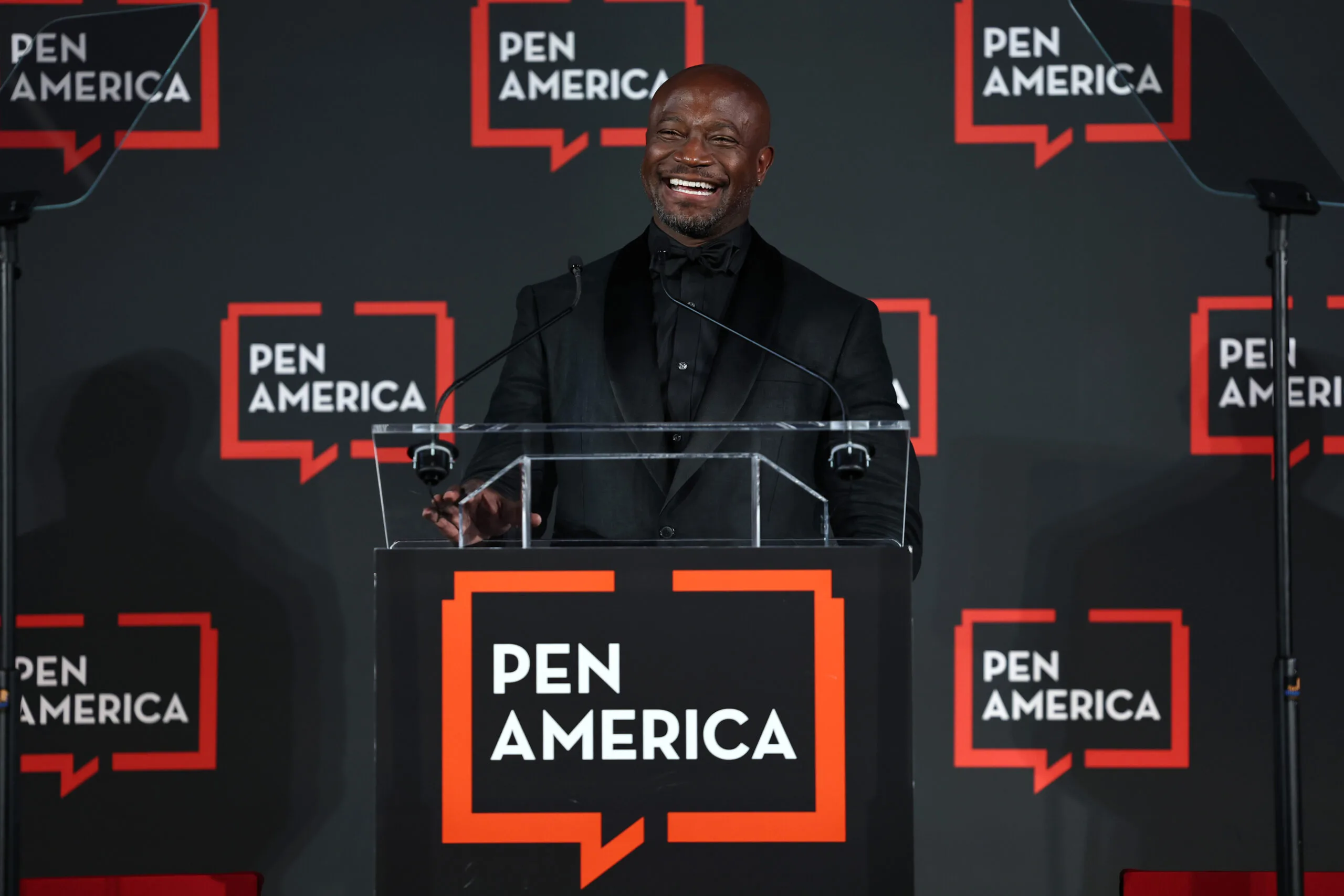 A man in a black suit smiles while speaking at a podium with a PEN America sign. The background features multiple PEN America logos on a dark backdrop.