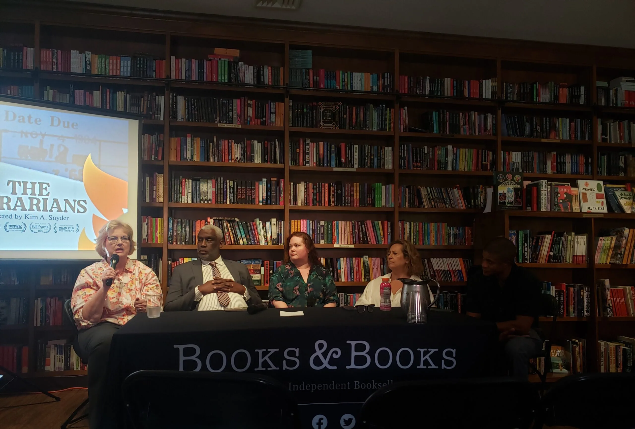 Five people sit at a panel discussion table labeled Books & Books in front of shelves filled with books. A screen displays The Librarians. One woman speaks into a microphone while others listen.